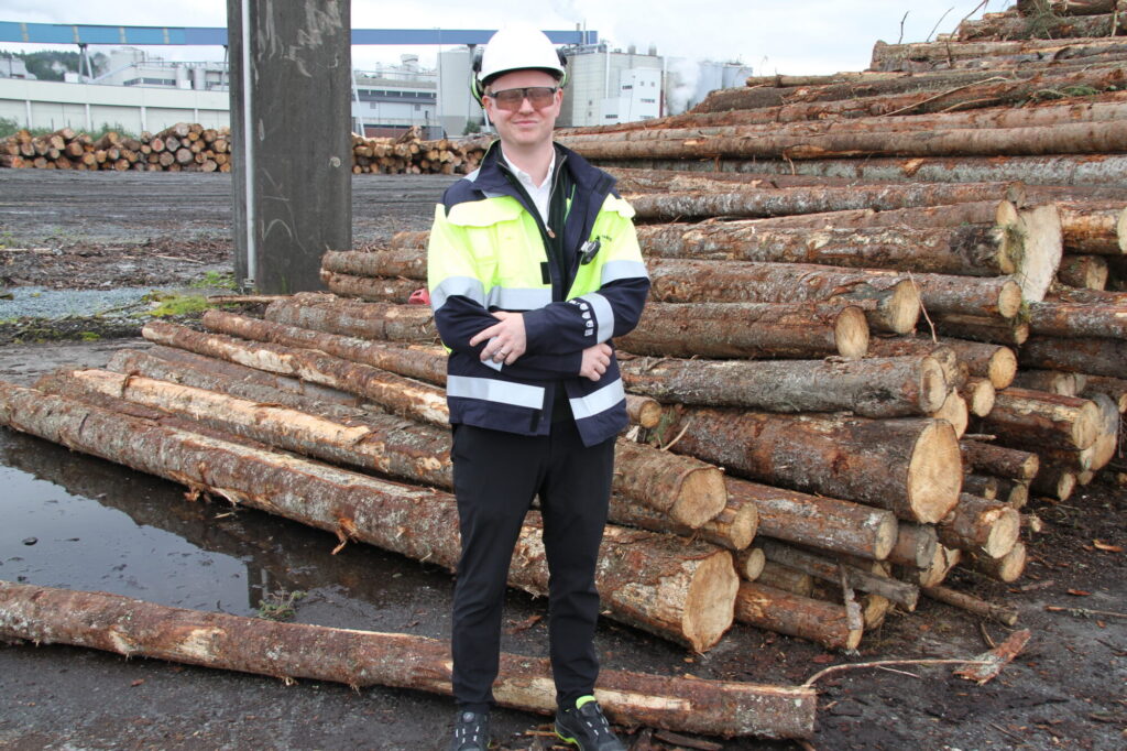 Mann i arbeidstøy og hjelm foran tømmerlunne på papirfabrikk.