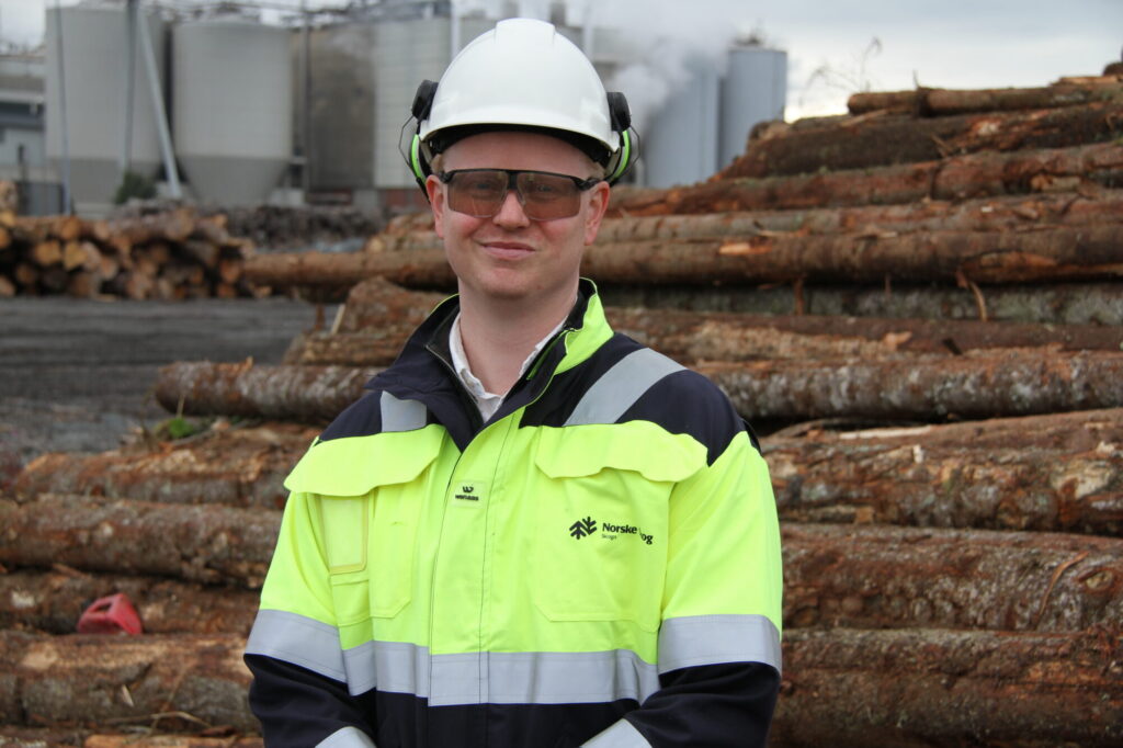 Man i arbeidstøy og hjelm på hodet foran tømmerlunne med papirfabrikk i bakgrunnen.