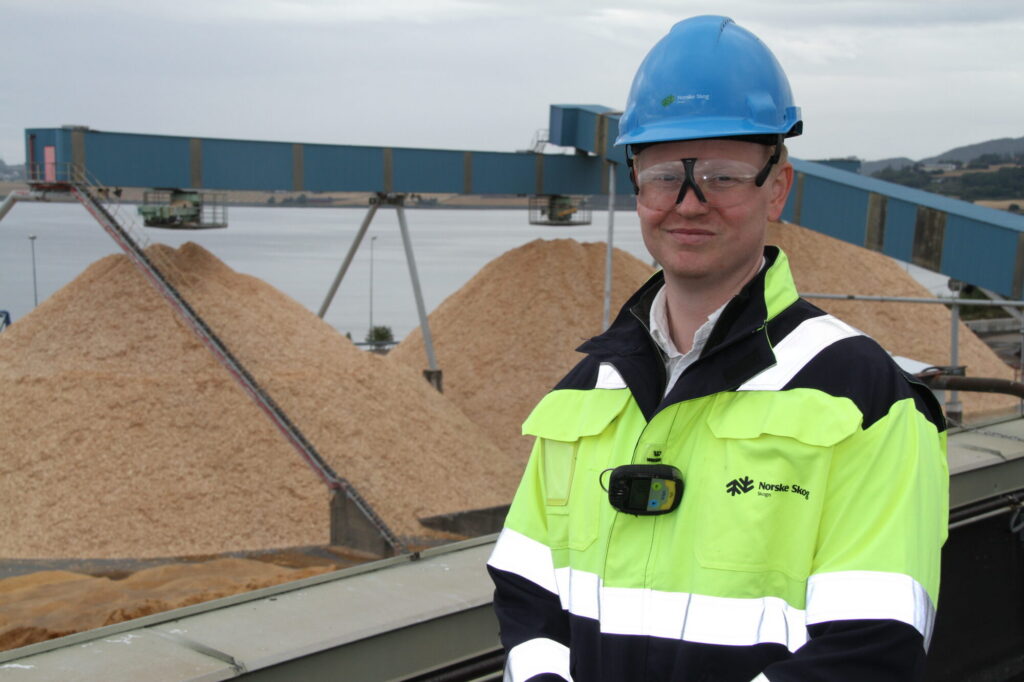 Mann i arbeidsklær med hjelm på hodet, foran stor treflishaug på papirfabrikk.