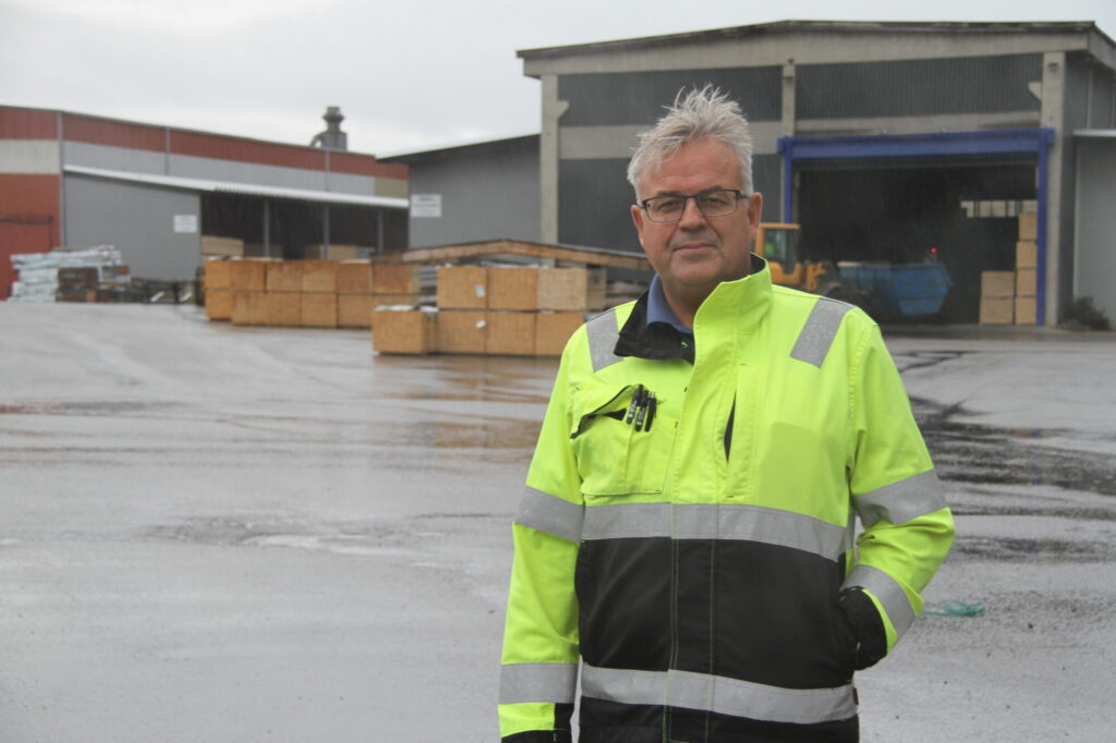 En man i arbeidsjakke foran materialstabler på et sagbruk.