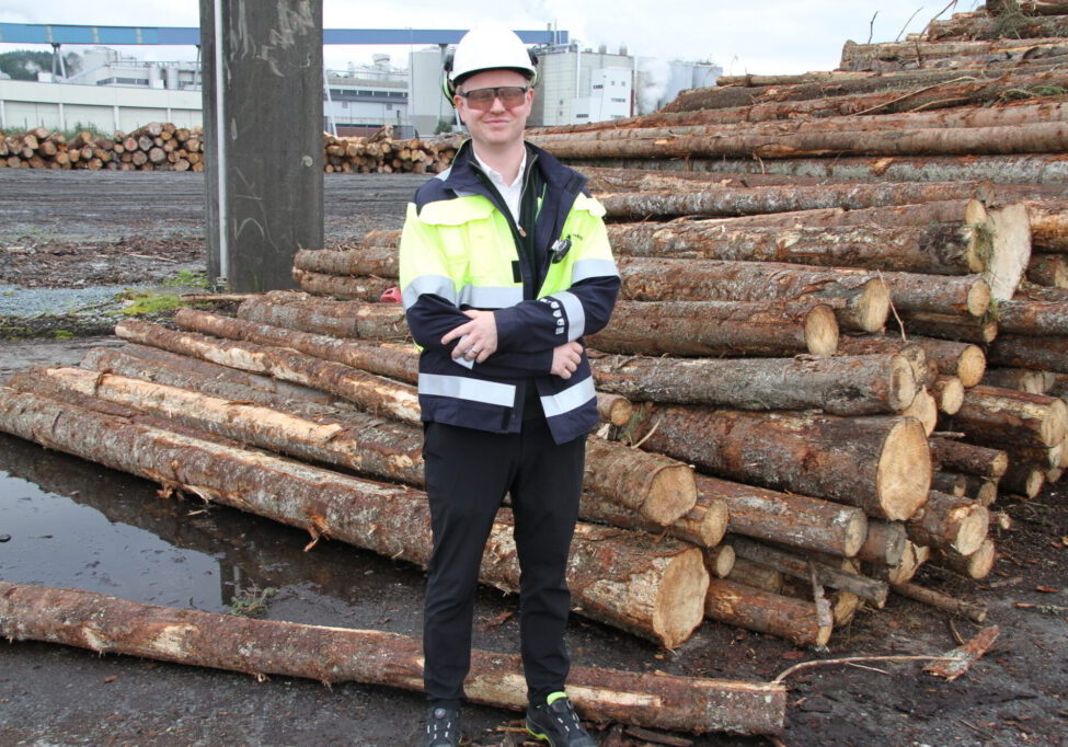 Mann i arbeidstøy og hjelm foran tømmerlunne på papirfabrikk.